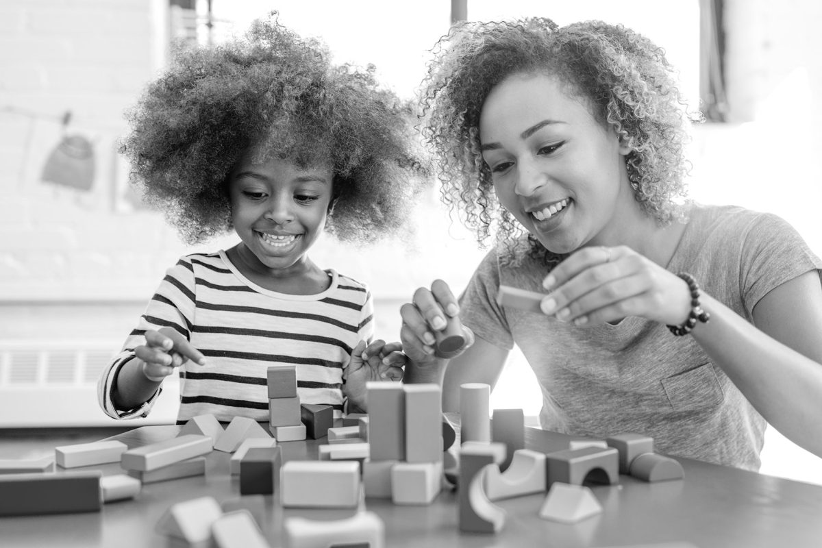 A child and a parent playing with blocks on a table in room with large glass windows.
